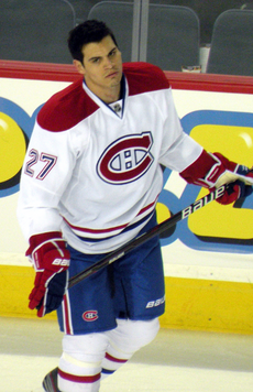 A hockey player with short dark hair looks to his right as he skates. He is in a white uniform with red and blue trim, the number 27 on his arms and a stylized "CH" logo on his chest.