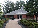 The Minnetonka Library, a one-story building with a large, triangular roof, out of which a triangular skylight arises. The building is situated among tall trees, and has a path leading away from its simple entrance. The path is adorned with bushes and a bench.