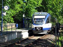 A blue-white three-segment diesel multiple unit on a single track, unelectrified railway line. It is stopping at a small station with one platform featuring a clock, lamps, rubbish bins and shelter, located in a forest.