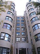 Detail of the entrance of a six-floor apartment building built from light-colored brick, with curved edges and windows at the corners and a grey concrete entryway.