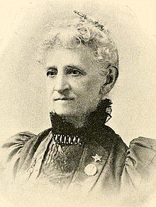 An older white woman, with white hair, wearing a high lace collar and large sleeves; she has a pin or medallion on her lapel.