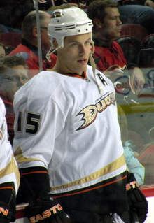 Upper body of a hockey player staring intently into the distance. He is in a white jersey with black, red and gold trim, with the word "DUCKS" scrawled across the chest.