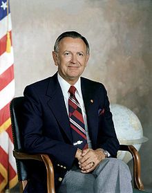 Formal portrait of a man in a jacket and tie seated in a chair in front of a U.S. flag.