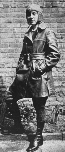 A young white woman dressed in a leather cap and thigh-lenght coat, wearing lettings and boots, standing in front of a brick wall with one leg bent at the knee