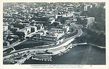 A road winds along the edge of a small cliff, with a mid-rise urban area behind it. Several prominent government buildings are located on both sides of the road. A bay of water is at the bottom of the cliff, where the Ottawa and Rideau Rivers meet.