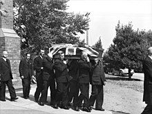 Black and white photo of a coffin which has been draped with a British flag being carried by pallbearers. The pallbearers are middle-aged men wearing formal suits, and three other middle aged men in suits are following the pallbearers.