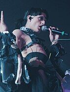 Arca sings into a microphone on stage. She has a light skin tone and black hair tied back in a ponytail, and is wearing a complicated black costume made up of straps, buckles, and zippered sleeves.