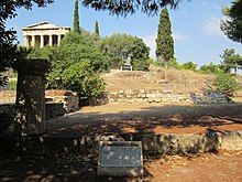 Color photograph of a ruin in the open. A single Greek column can be seen in the foreground on top of a compound wall. In the background are some trees, and at the rear left is a Classical Greek building.