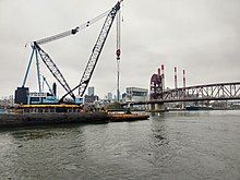 A large crane on a barge, about to lift the triangular metal frame supporting 3 turbines