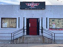 Grey stcco building with red door. Eagle shaped sign above door reads "Denver Eagle"