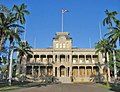 Photograph of the front of ʻIolani Palace, flanked by palm trees with the Hawaiian flag flying atop the central tower.