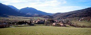 A small village in a green field stands before a low, blue mountain peak