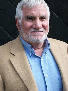 Portrait of Henry Marsh, caucasian man in his mid-60s at the time of photograph, with white short hair and slight beard, smiling and looking toward the camera, wearing a blue button down collared shirt and tan jacket.