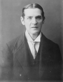 A black and white portrait of a formally dressed individual with neatly combed hair and a serious expression.