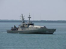 A grey warship underway. Land is visible in the background.