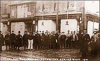 A sepia-toned photograph shows a group of men standing in front of a row of buildings with signs indicating shops or businesses. The men are dressed in early 20th-century attire, including hats and overcoats. The image is labeled "Tonypandy after the strike riot 1910”. The men appear somber, and the buildings show signs of disrepair or damage.