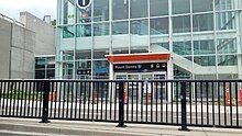 Photo of a glass station building with an entryway at ground level. An elevator and staircase are visible inside.