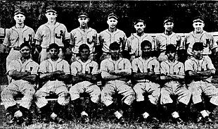 A black and white photo of 14 in two rows (7 standing, 7 sitting in chairs) wearing light baseball uniforms, each with a "J" on the chest