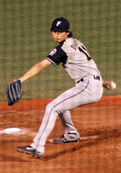 A young Japanese man wearing a grey baseball uniform takes a step forward as he begins to throw a baseball.