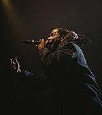 A black man, dressed in all black, stands side on to the camera. He is performing on stage with a microphone in his hand