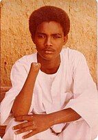 shown is Abdul Rahman Ali, a young Sudanese dressed in a jalabiya, a traditional Sudanese white garment, sitting against a textured, warm-toned wall. Ali has a calm and contemplative expression, resting their left arm on their lap while their hand supports their face. Ali's right arm is amputated just below the elbow.