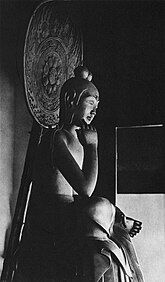 Three-quarter view of a seated statue in half-lotus position. The right foot rests on the left upper leg, the right elbow rests on the right knee with the right hand close to the head. The hair of the statue is sculpted with two top-knots. There is a halo behind the statue's head. Black and white picture.