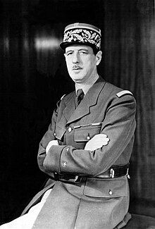 Middle-aged Caucasian man with a long face and small mustache in a half-profile, half-portrait pose, with his arms folded over his chest. He has a very prominent nose and is wearing a ceremonial military uniform with a tie, suit and a cylindrical military hat.