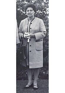 Woman with dark hair and glasses with handbag in white coat and black shoes.