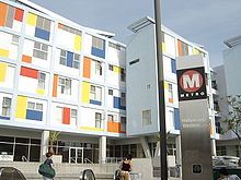 A silver pillar indicates the entrance to a subway station. A multicolored building is located in the background.