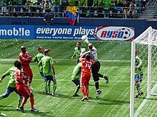 Several players in red uniforms and green uniforms in front of goal with a goalkeeper in black leaping with his hand hitting a soccer ball