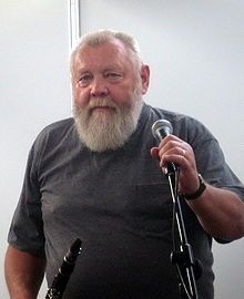 Ivo Pešák wearing a grey t-shirt with breast pocket, holding onto a microphone in a stand with one hand, with a clarinet in the other, looking directly at camera