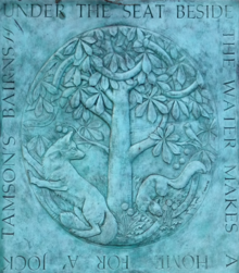 Copper plaque with patina depicting two foxes under a tree. Around the outside is the inscription: "Under the seat beside the water makes a home for a' Jock Tamson's bairns"