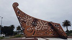 An elaborate Māori carving painted reddish brown carved like the end of a waka canoe set into concrete