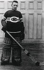 An ice hockey Goaltender stands, also wearing a Canadians jersey, small leg pads and old style gloves, he also is without a mask, while posing for the photo but it was also the style of the time.