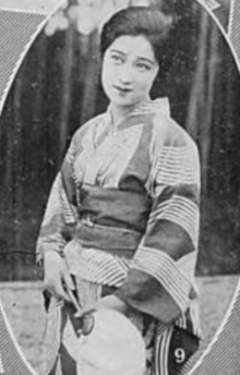 A Japanese woman photographed outdoors, wearing a striped kimono and holding a fan