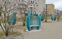 Blue sculptural egg-shaped arms supporting a seat, surrounded by small trees in front of a group of apartment houses