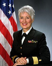 Older white woman with short grey hair, smiling, in United States Navy uniform, with United States flag in background.