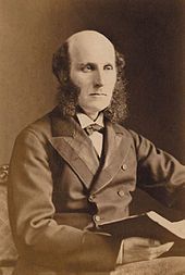 A well-dressed middle-aged man with a bald head and bushy sideburns, wearing a dark suit and carrying a large book