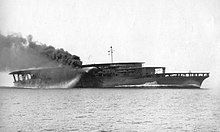 An aircraft carrier steaming at full speed. Thick black smoke billows from its curved smoke stack.