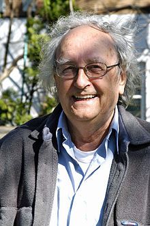 Laurent Lachance wearing a white t-shirt, a light-coloured shirt, and a grey fleece jacket, standing outdoors with lightly windblown hair, grinning at camera