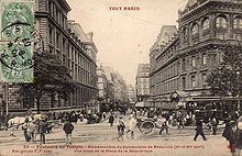 A sepia photograph looking down a built-up street, with the tramway to the right. A small crowd waits to board the tram.