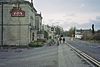 Gray stone building on the left with a pub sign outside it. A road is central to the picture with a white coloured building on the right.