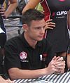 A man in his early twenties looking to someone outside the camera. He is wearing a black T-shirt with sponsors logos and his holding both his hands together/