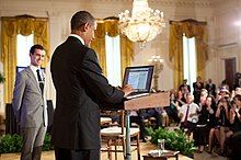 Obama in blue suit standing at a podium in front of an audience as a man in a light grey suit looks on.