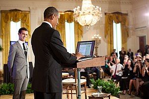 Black man in blue suit standing at a podium in front of an audience as a white man in a light grey suit looks on.