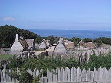 A color picture of the reconstructed Plymouth Plantation in Plymouth. All upright wood logs are faded pale from the sun.