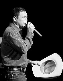 A black and white photo of Pulido wearing a dress shirt tucked in jeans with his cowboy hat on one hand and a microphone on the other. A Keyboardist and his instrument can be seen in the foreground of the photo.