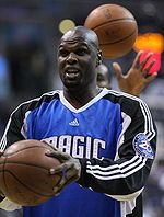 A dark-skinned black man wearing basketball warm-up gear holds a basketball in front of him while making a surprised expression.