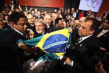 A man holds a flag surrounded by several people celebrating. In the background, photographers record the moment.