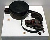 Black disc-shaped landmine on a white display stand, alongside broken fragments of another landmine.